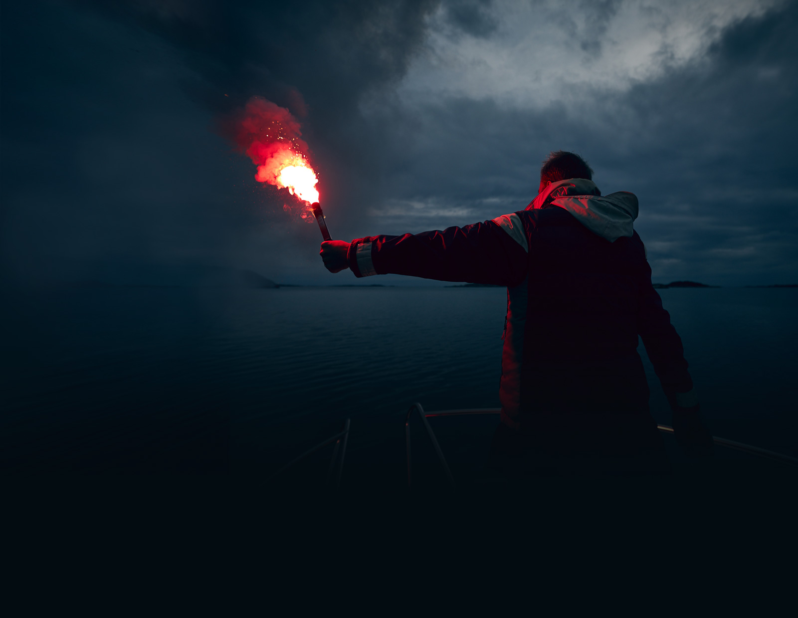 Un hombre situado en la proa de un barco. Sostiene en la oscuridad uno de los productos de IKAROS: la bengala de mano roja. La luz roja procedente de la bengala de socorro ilumina la oscuridad.