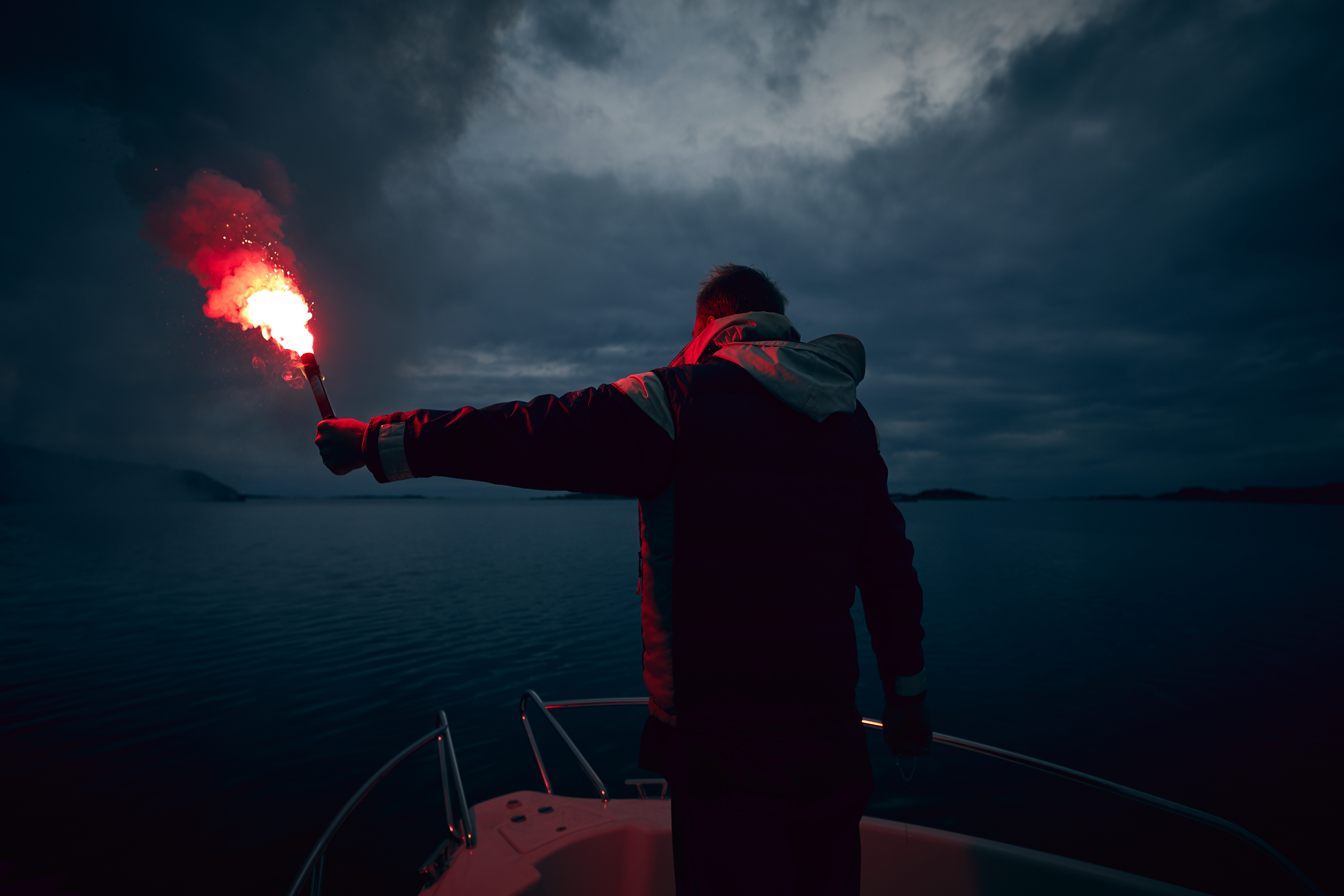 A man stands in the bow of a leisure boat. It's dark outside and he is holding one of IKAROS products, the red handheld flare. The red smoke coming from the emergency flare lights up the dark.