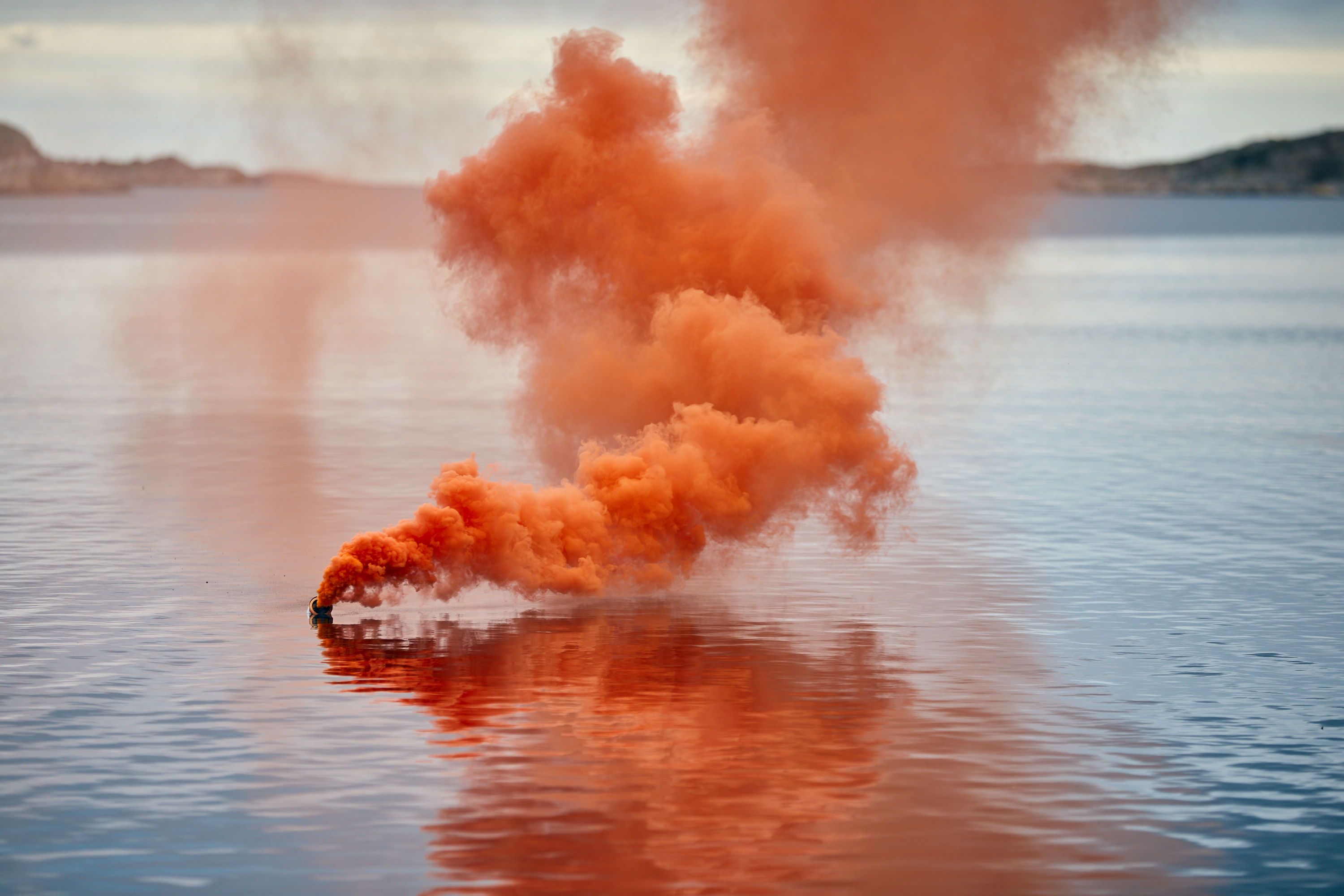 A buoyant distress signal floating at sea in daylight, releasing a strong colored smoke spreading over the water, visible from kilometers away.