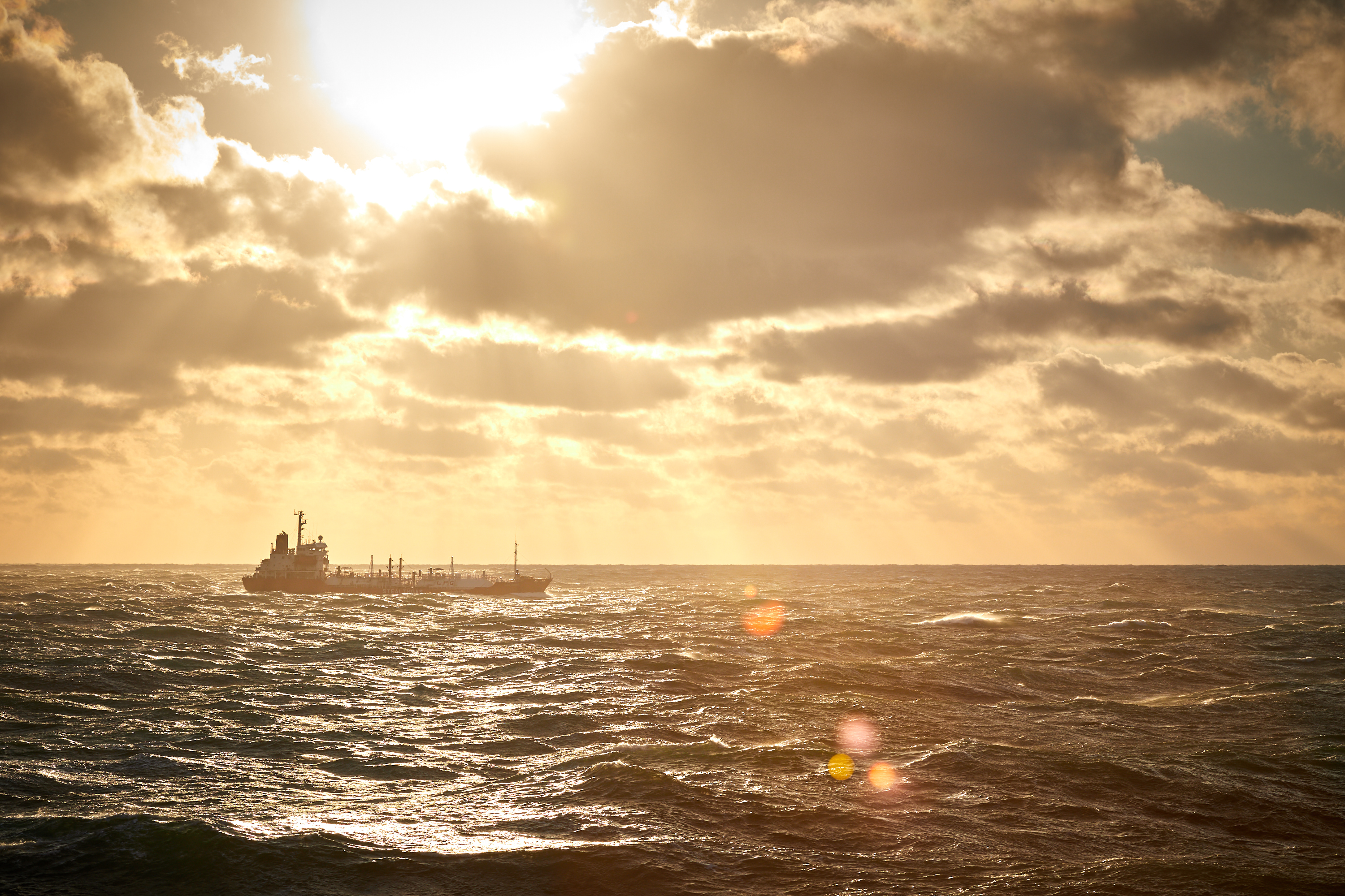 A lonely cargo vessel on an open sea. The sky is dramatic and the sun shines through the clouds. No signal flares are visible.
