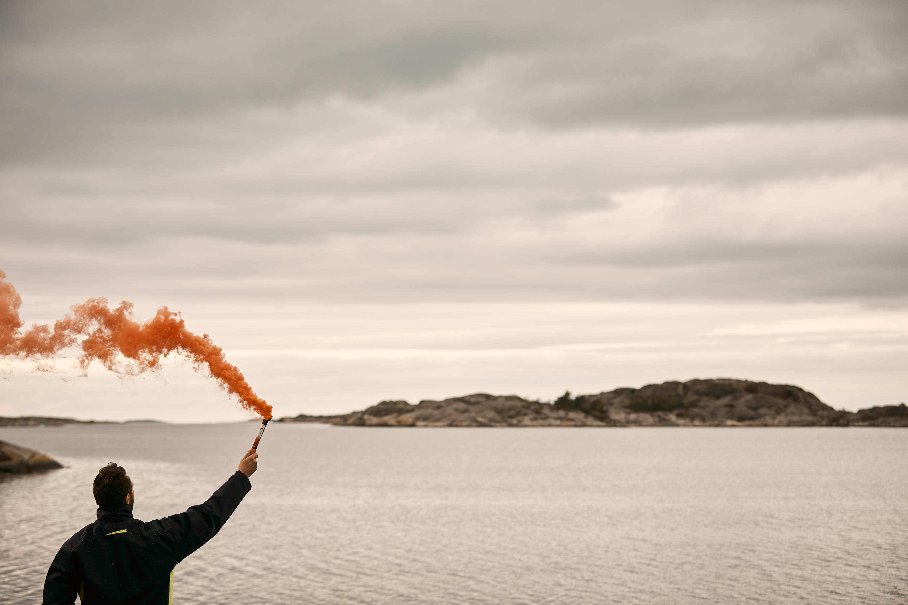 A man stands on a shore, holding one of IKAROS signal flares, the orange handsmoke. The orange smoke coming from the distress signal spreads in the wind.